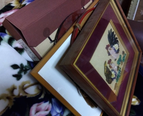鶏が描かれた額装の貼り絵作品と他の額縁、保管箱が布団の上に重ねて置かれた様子｜生前整理中の骨董品