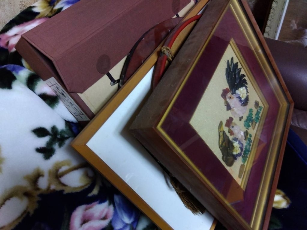 鶏が描かれた額装の貼り絵作品と他の額縁、保管箱が布団の上に重ねて置かれた様子｜生前整理中の骨董品